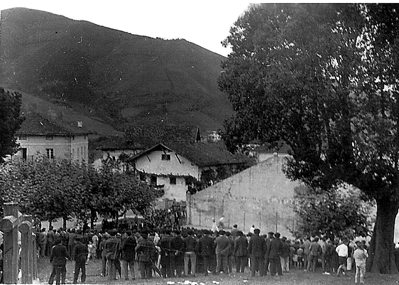 Urrunetik atarata, Berriatuko frontoi zaharrean, esku pilota partida ikusten hainbat herritar. [Argazkia]